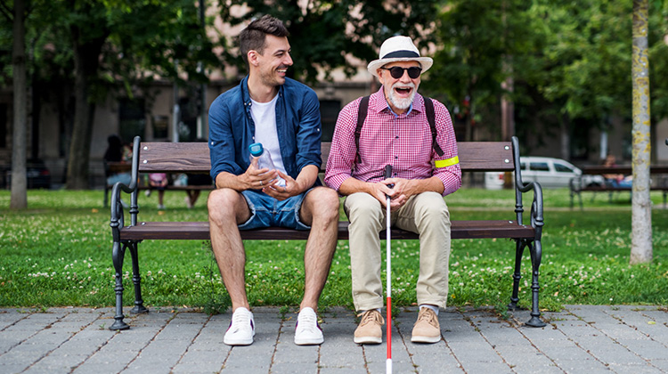 Alter blinder Mann auf einer Parkbank lacht zusammen mit einem jungen Mann